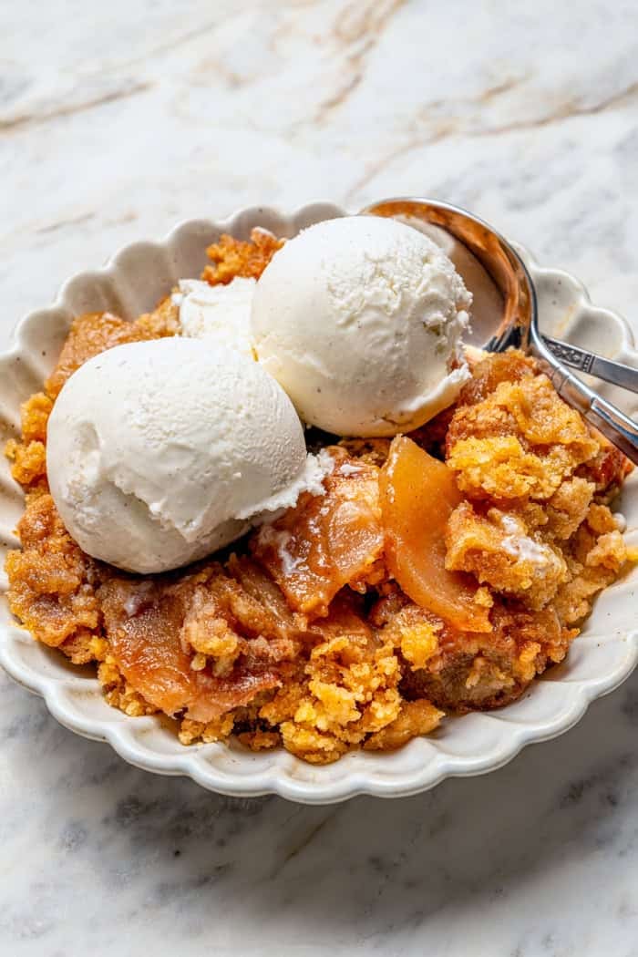 an individual serving of apple dump cake with scoops of vanilla ice cream, in a white dessert dish