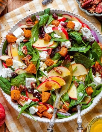 overhead photo of a wood table with a platter full of fall harvest salad with fresh greens, butternut squash, fresh greens, pecans, cranberries, and goat cheesegreens, pecans, cranberries, and goat cheese