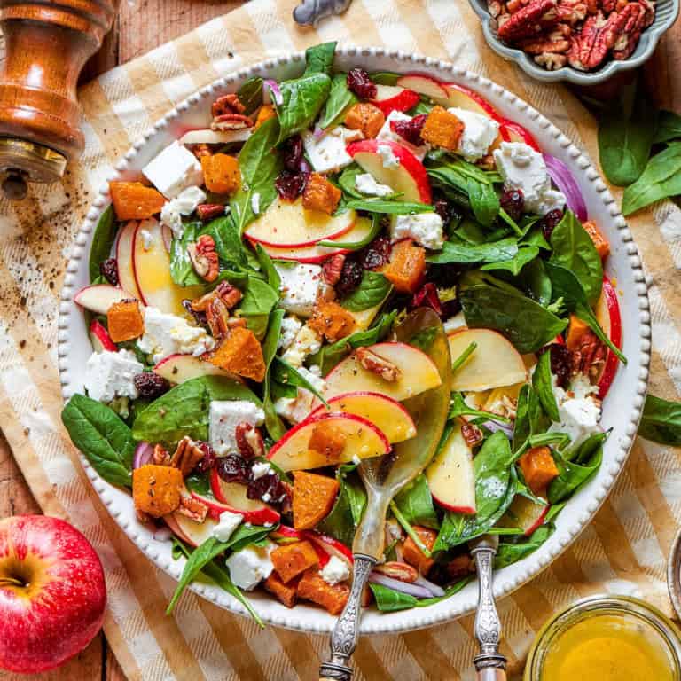 overhead photo of a wood table with a platter full of fall harvest salad with fresh greens, butternut squash, fresh greens, pecans, cranberries, and goat cheesegreens, pecans, cranberries, and goat cheese