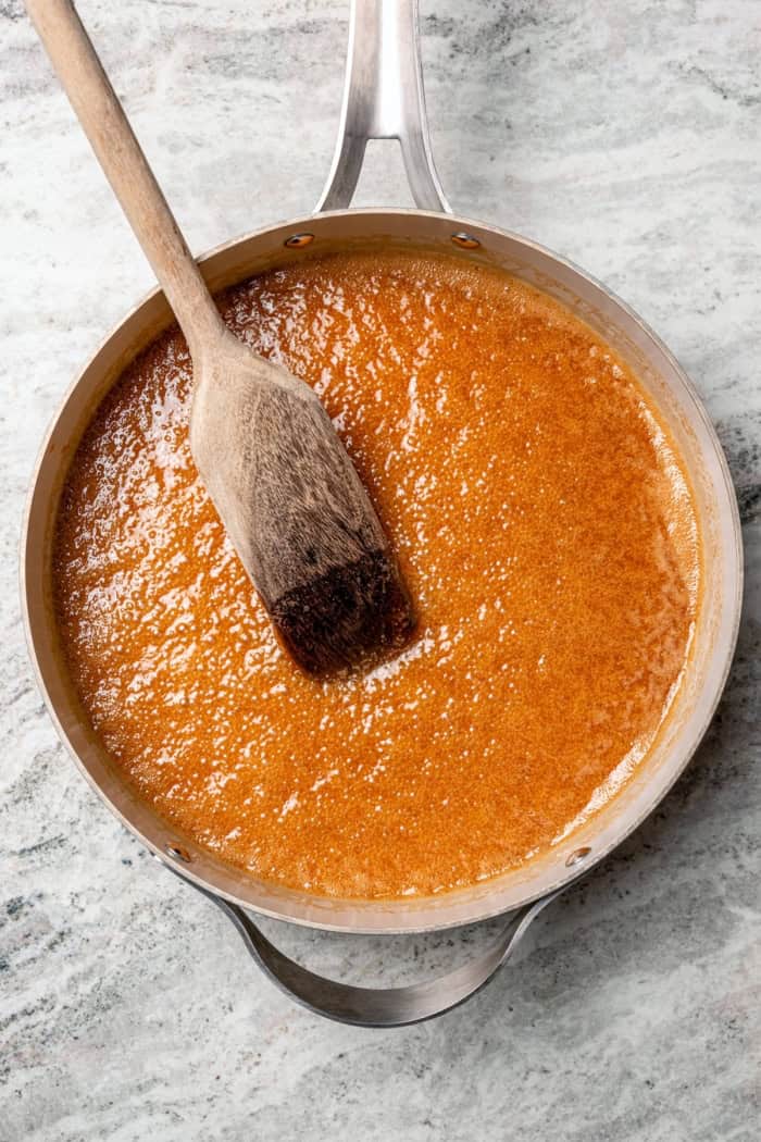 These Pecan Pie Bars have a caramel filling, buttery shortbread crust and NO corn syrup! Honey and brown sugar adds the perfect sweetness. Caramel for pecan pie bars in saucepan with wooden spoon.