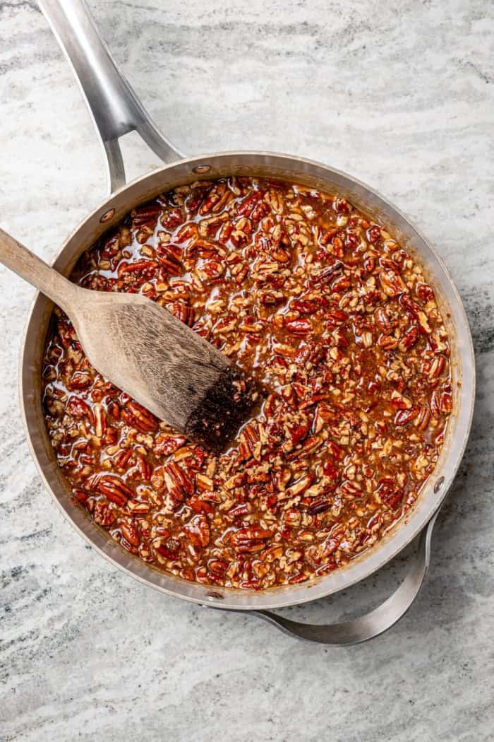 These Pecan Pie Bars have a caramel filling, buttery shortbread crust and NO corn syrup! Honey and brown sugar adds the perfect sweetness. Pecan pie bar filling in pan with wooden spoon.