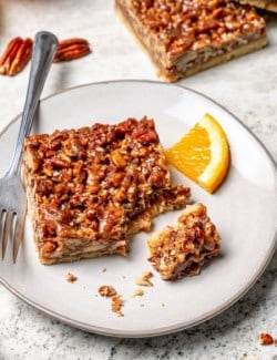 Pecan pie bar on plate with fork and orange slice.