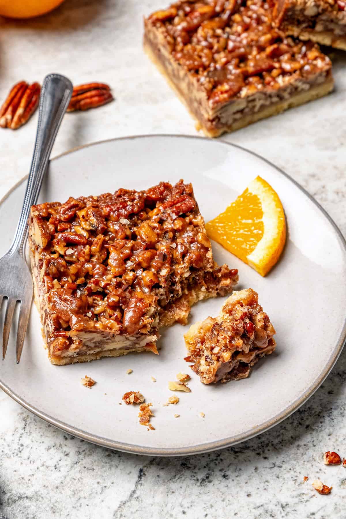 These Pecan Pie Bars have a caramel filling, buttery shortbread crust and NO corn syrup! Honey and brown sugar adds the perfect sweetness. Pecan pie bar on plate with fork and orange slice.