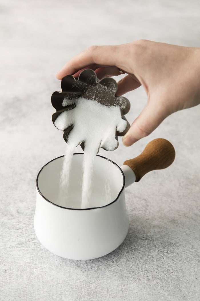a hand pouring sugar from a small tin into a small white pan