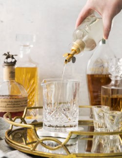 a hand pouring simple syrup from a clear bottle into a cocktail mixing glass