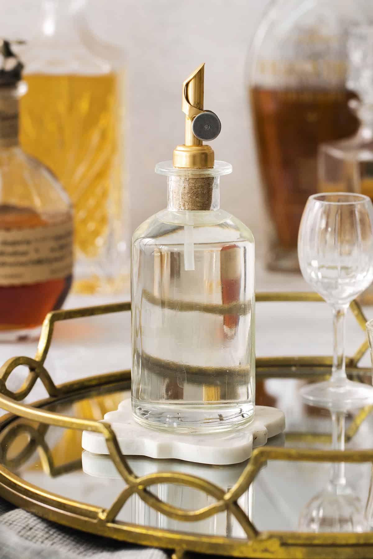 a clear bottle with a gold pour spout, full of simple syrup, sitting on a gold mirrored cocktail tray