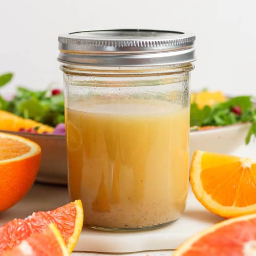 a jar of citrus vinaigrette, with cut fresh citrus and a bowl of salad greens