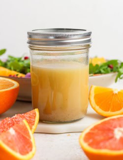 a small jar of citrus vinaigrette salad dressing, with cut fresh citrus and a bowl of salad greens