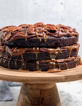 a layered chocolate, caramel, and pecans Turtle Cake on a wooden cake pedestal