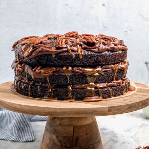 a layered chocolate, caramel, and pecans Turtle Cake on a wooden cake pedestal