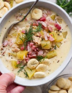 a hand holding a white bowl of clam chowder with pork belly