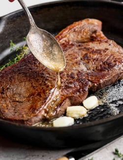 Spooning garlic butter over steak in cast iron skillet.