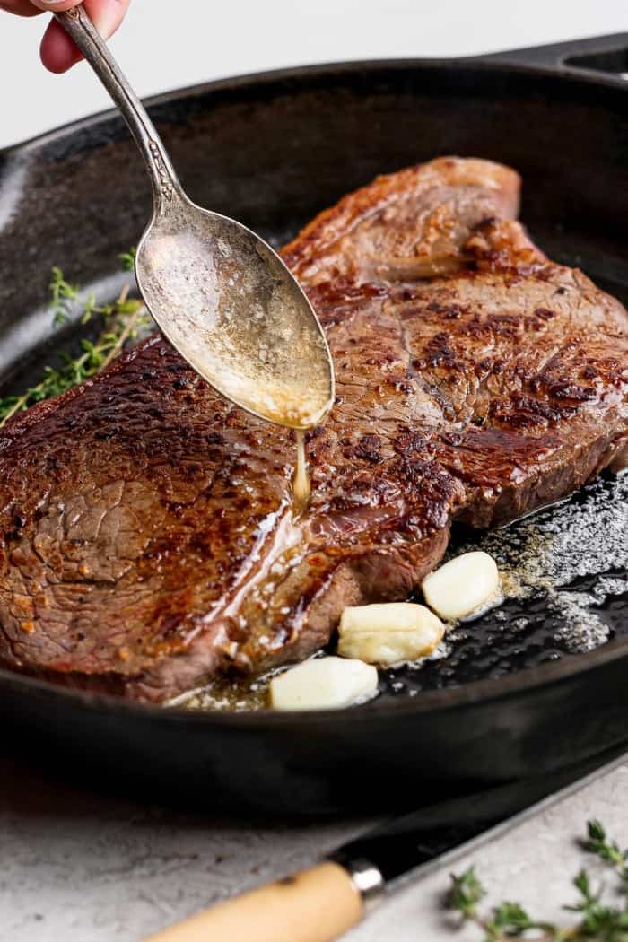 Spooning garlic butter over steak in cast iron skillet.