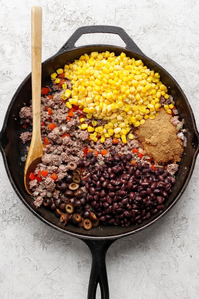 overhead photo of ingredients separated in a cast iron skillet: cooked ground beef, corn kernels, taco seasoning, black beans, black olives