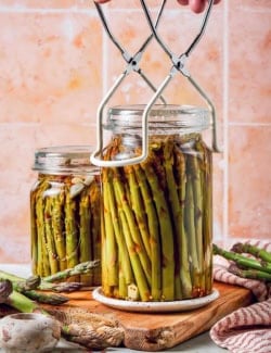 a hand setting a jar of pickled asparagus down onto a wood cutting board, using a jar lifter tool