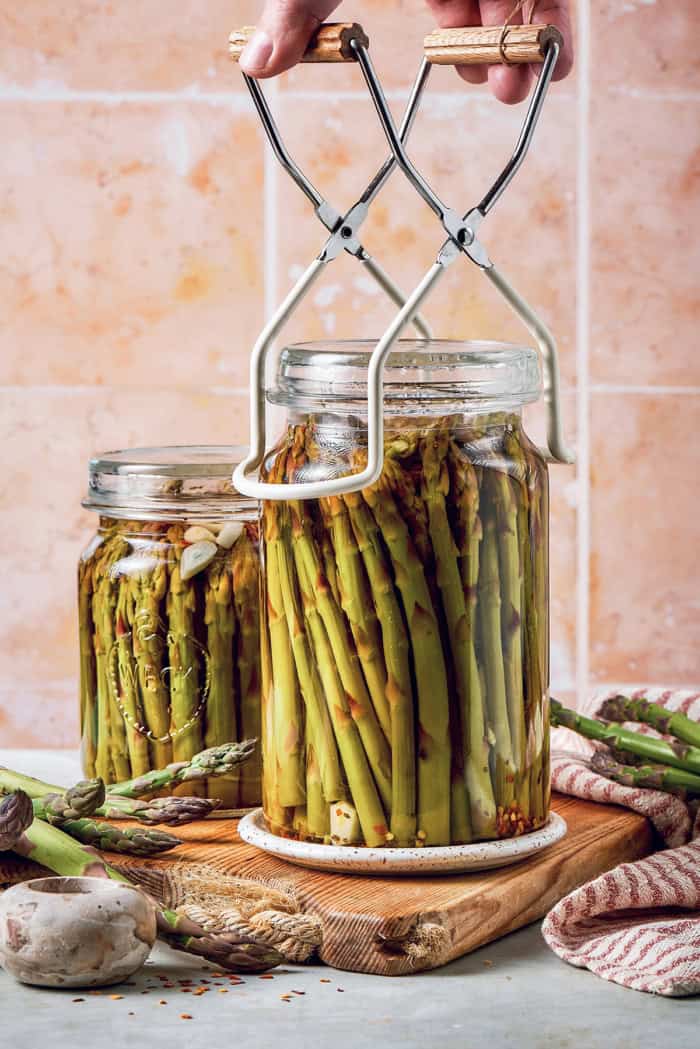 a hand setting a jar of pickled asparagus down onto a wood cutting board, using a jar lifter tool