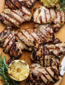 Grilled lemon chicken thighs on cutting board with fresh herbs and charred lemon halves.