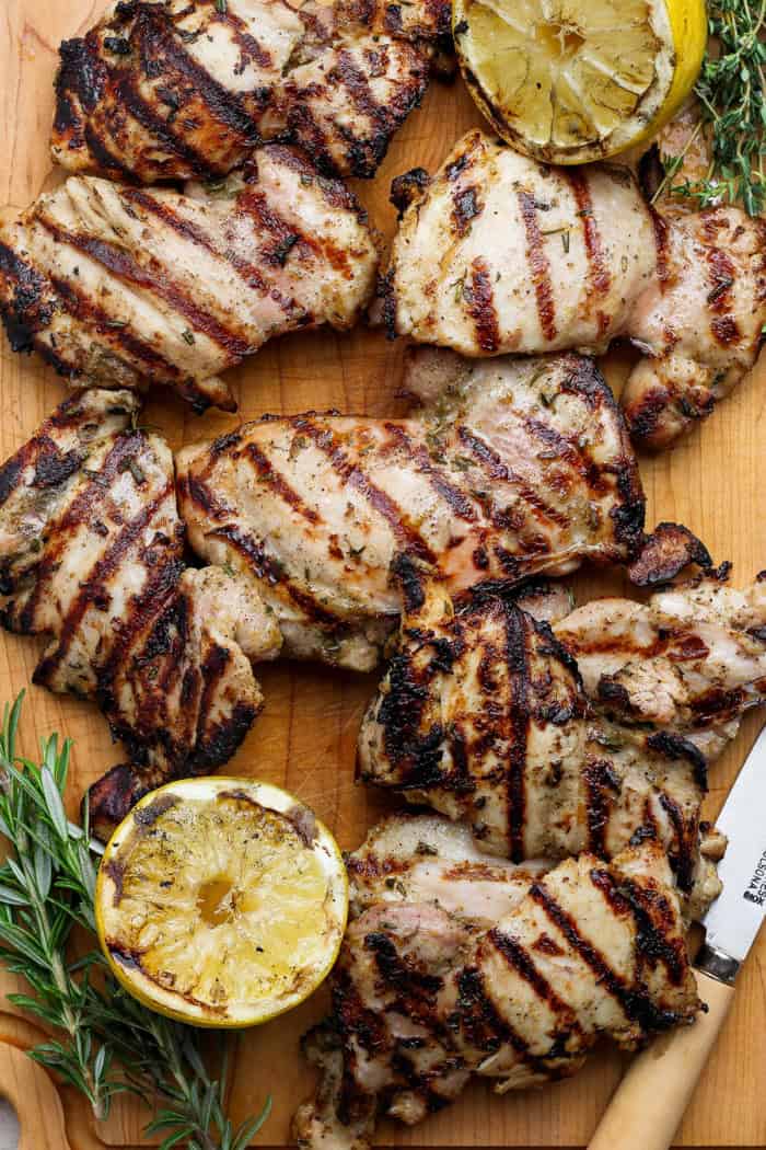 Grilled lemon chicken thighs on cutting board with fresh herbs and charred lemon halves.