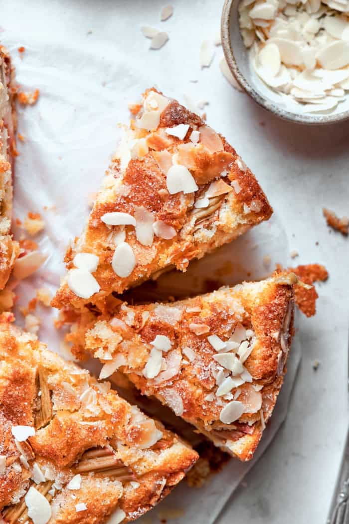 This simple Norwegian rhubarb cake is tender and delicate, with jammy rhubarb and a sprinkling of sliced almonds and cinnamon sugar on top. Two slices of Norwegian rhubarb cake pulled away from rest of cake.