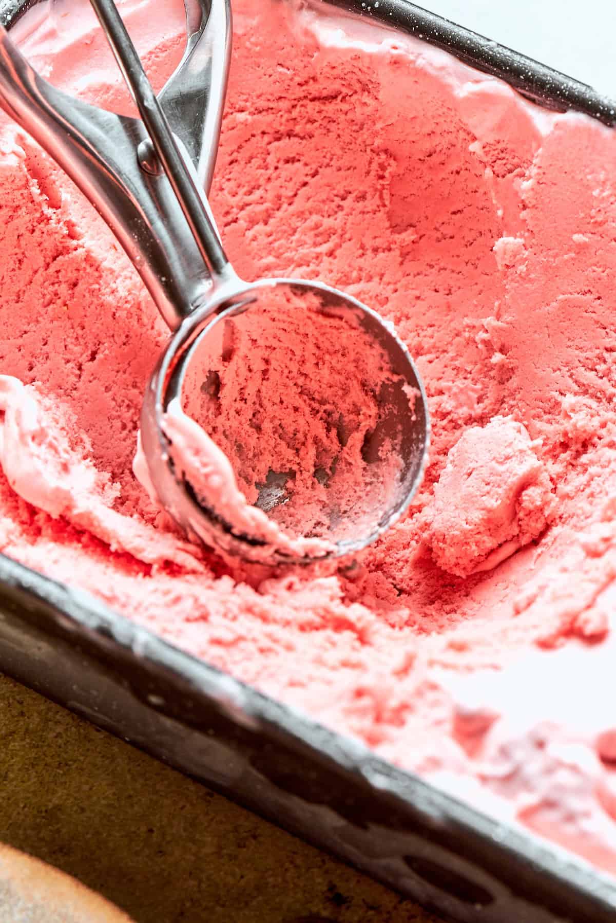 This homemade rhubarb ice cream is ultra creamy, with tart rhubarb flavor and a lovely blush-pink color. Such a fun summer dessert! Ice cream scoop in loaf pan of rhubarb ice cream.