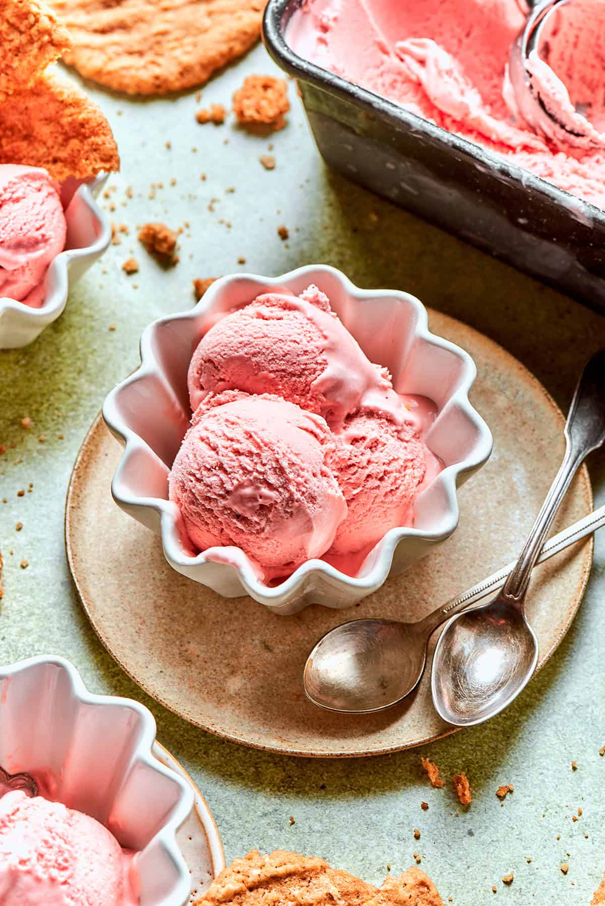 This homemade rhubarb ice cream is ultra creamy, with tart rhubarb flavor and a lovely blush-pink color. Such a fun summer dessert! scoops of pink rhubarb ice cream in white fluted bowls, with crispy oat cookies