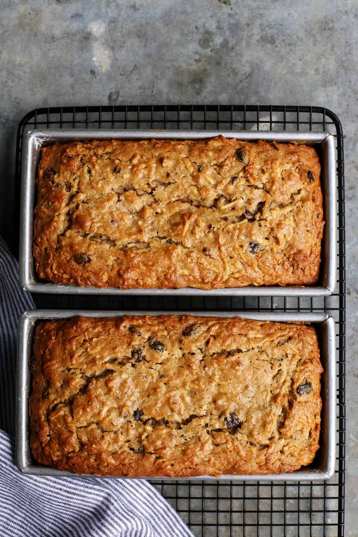 overhead photo of two baked loaves of apple bread in loaf pans, on a wire cooling rack