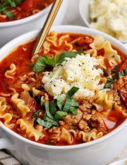 close-up photo of lasagna soup with dollop of cheesy yum and fresh basil on top, in white bowl