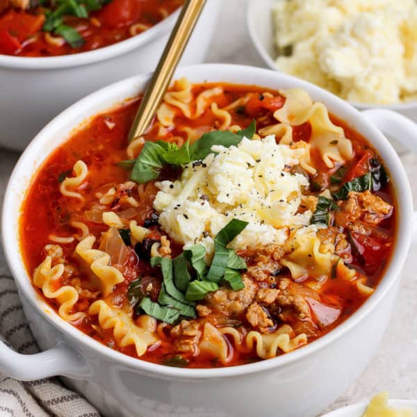 close-up photo of lasagna soup with dollop of cheesy yum and fresh basil on top, in white bowl
