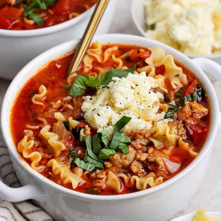 close-up photo of lasagna soup with dollop of cheesy yum and fresh basil on top, in white bowl