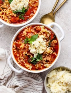 overhead photo of two white bowls filled with lasagna soup and topped with dollops of cheesy yum and fresh basil