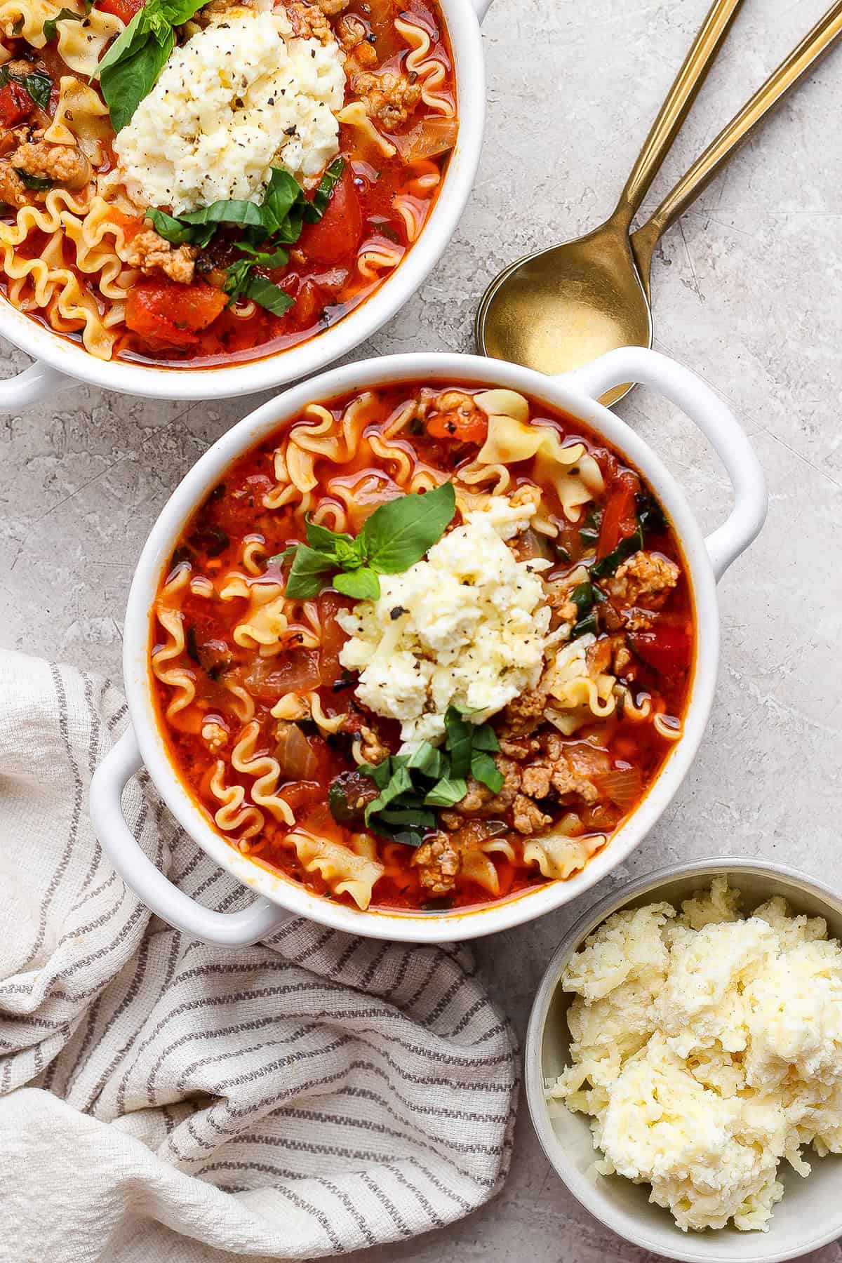 overhead photo of two white bowls filled with lasagna soup and topped with dollops of cheesy yum and fresh basil
