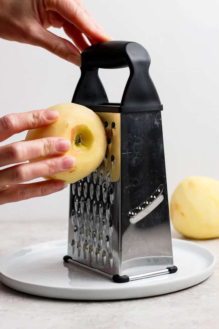 hands holding a peeled whole apple and shredding it with a box grater