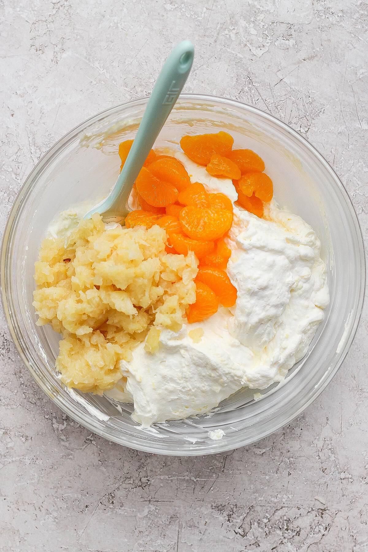 adding crushed pineapple and mandarin oranges to a fluffy mixture of Cool Whip, buttermilk, and vanilla instant pudding mix in a large clear bowl
